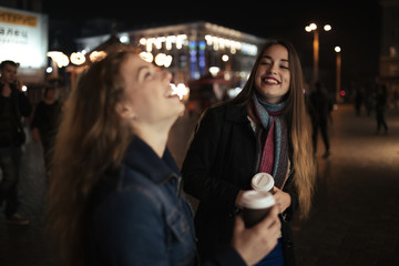 Two women friends walking in city street at night and drinking coffee