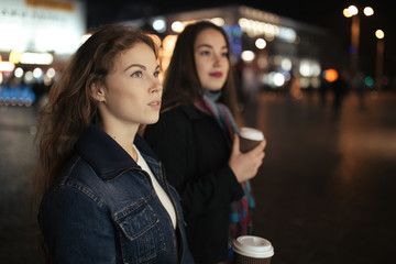 Two women friends walking in city street and drinking coffee at night