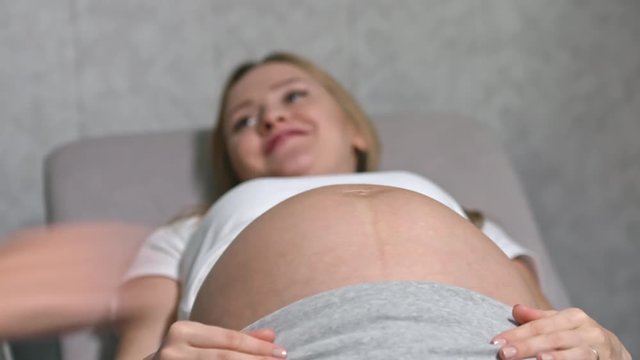 Hands of female doctor measuring belly of smiling pregnant woman lying on examination table, then writing down information