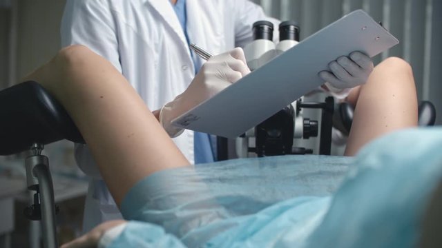 Over the shoulder shot of woman lying on gynecological chair and being examined by female doctor with colposcopy microscope 