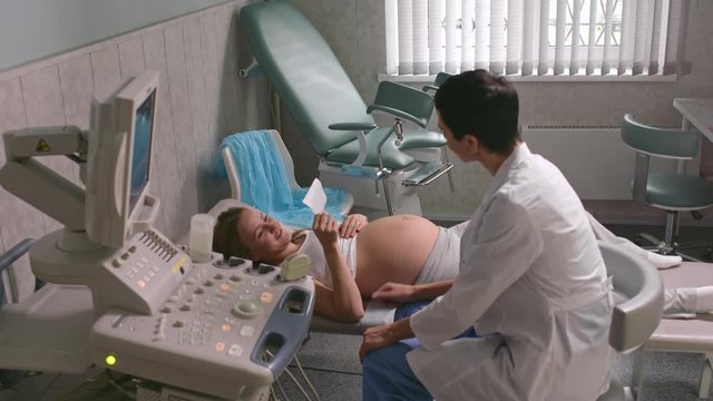 Happy pregnant woman lying on examination table and looking at female doctor giving her ultrasound scan picture