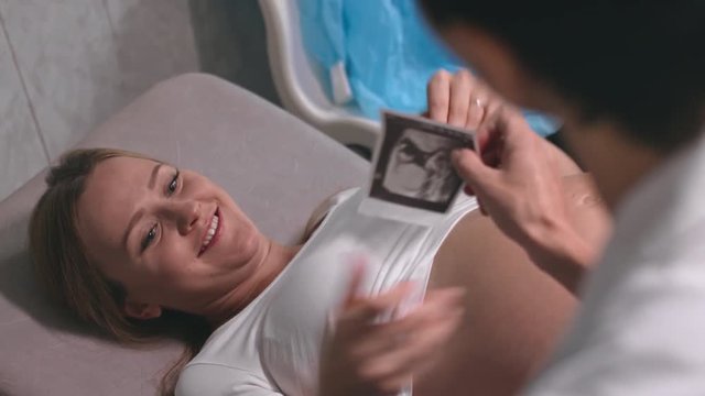 Over the shoulder shot of female doctor stroking belly of happy pregnant patient and giving her ultrasound scan picture of baby