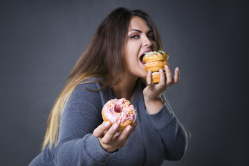 Happy beautiful young caucasian plus size model posing with donuts on a gray studio background