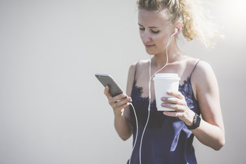 Girl with blond hair,wearing blue top,standing and using smartphone while listening to music on headphones.In her hand she holds paper cup with drink.In the background a white wall.Girl using gadget.