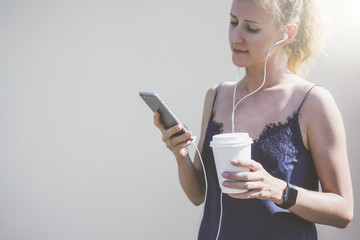 Sunny summer day,young woman dressed in blue silk top,standing near a white wall and listening to music on headphones.In one hand she holds smartphone in other-cup with a drink.Girl using gadget.