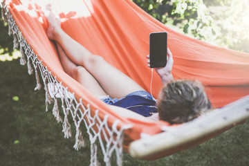 View from behind,young woman in shorts listening to music while lying in hammock and holding a smartphone with a blank screen.Selective focus,film effect,blurred foreground,mock up.Girl using gadget.