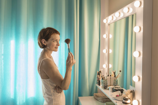 Woman holding make-up brush standing in front of mirror at dressing table