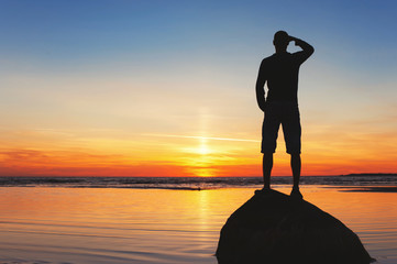 Young man silhouette standing on the rock at sunset background