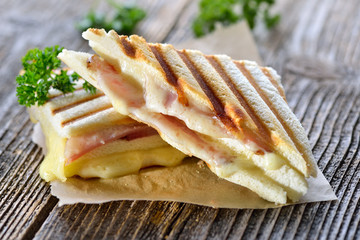 Getoastetes und im Kontaktgrill gepresstes italienisches Panini mit Schinken und Käse - Pressed and toasted double panini with ham and cheese served on sandwich paper on a wooden table