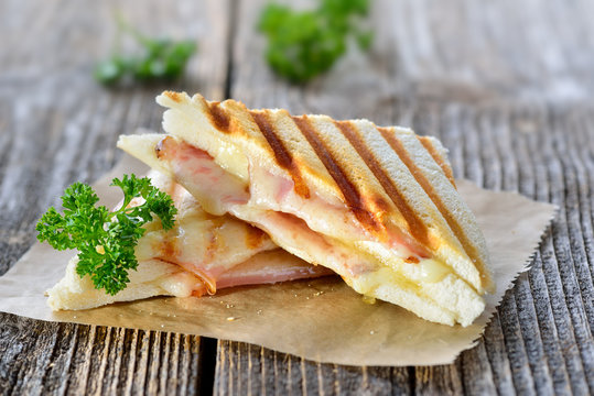 Getoastetes und im Kontaktgrill gepresstes italienisches Panini mit Schinken und Käse - Pressed and toasted double panini with ham and cheese served on sandwich paper on a wooden table