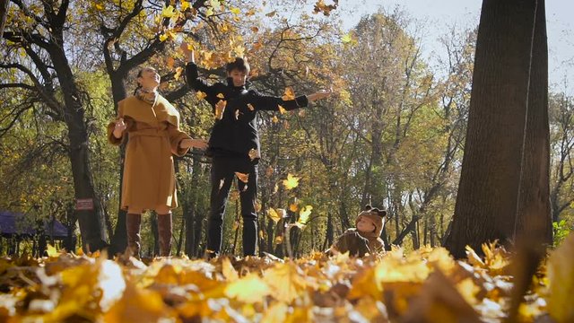 Happy family playing with autumn leaves in park. Mom and dad throw leas up in the air. Small child look at them and having fun.