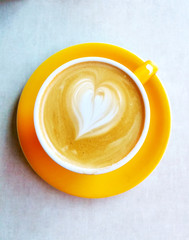 Top view of a heart shaped in a yellow cup of latte, cappuccino coffee. Cafe art with milk, cofee, round ceramic glass and plate for coffe lovers - aka latte art.
