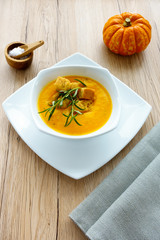 Pumpkin soup with herbs, sea salt flakes in wooden bowl and a munchkin pumpkin on light brown wooden background. 