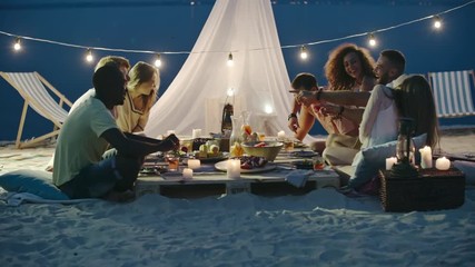 Young friends sitting at the beach picnic at night, talking, laughing, toasting and drinking summer cocktails - Powered by Adobe
