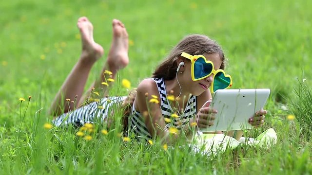 Beautiful girl in big sunglasses with tablet pc lies on green grass. Little girl in glasses lies on the green grass and uses tablet computer. Girl playing the game on his tablet computer