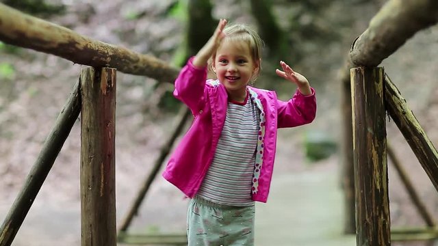 Little girl stands on a wooden bridge and does dance moves. Funny little girl in pink jacket on the nature