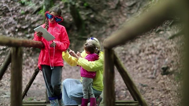 Mother and two daughters with smartphone and tablet computer on the nature. Family with electronic devices on the wooden bridge in the forest. Women in color windcheaters with phone and tablet pc