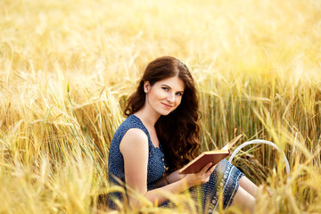 Portrait of cute pretty happy young woman with basket in dress reading book sitting on a grass in field. Sunny morning. harvest season, autumn nature, healthy concept