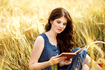 Portrait of cute pretty happy young woman with basket in dress reading book sitting on a grass in...