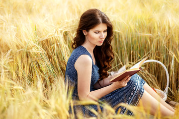 Portrait of cute pretty happy young woman with basket in dress reading book sitting on a grass in field. Sunny morning. harvest season, autumn nature, healthy concept