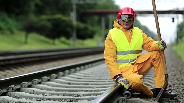 Railway worker in yellow uniform with shovel in hand sits on railway line. Railwayman in red hard hat sits on rail and looks at the camera. Workman with spade on railway track. Railway construction