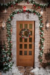 Winter wreath hanging on a door of house decorated by Christmas pine branch with red baubles and decorative snow. New Year tree standing near brick wall.
