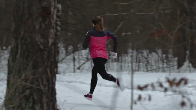 Tracking shot of athletic young woman running in snow through forest during winter cross-country training