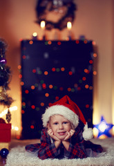 Pretty small girl wearing red Santa hat lying on warm carpet over red lights background, Concept of Christmas holidays and celebration. 