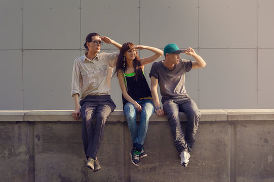 Three Friends Are Sitting On The Fence And Looking Into The Distance. Urban Sunset Lifestyle. Two Men And Woman Are Watching The Sunset