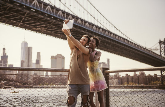 Multi-ethnic couple taking selfie through tablet computer against Manhattan Bridge and city skyline