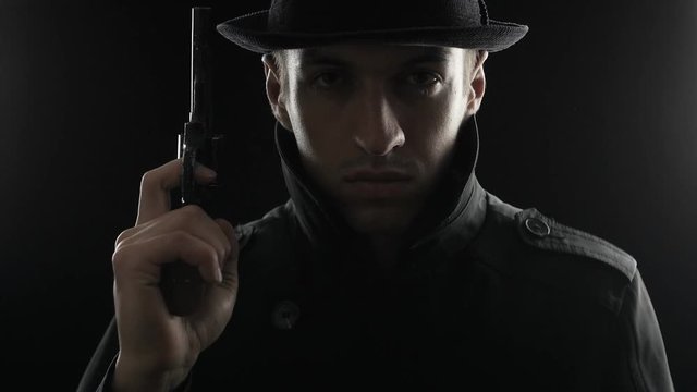 Portrait of a mafioso in a hat and a black cloak with a gun in hand