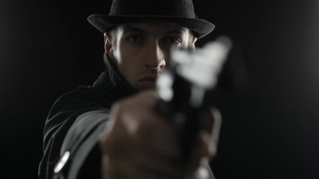Portrait of a gangster in a hat and a black cloak threatens with weapon. Mafioso aiming a gun at the camera