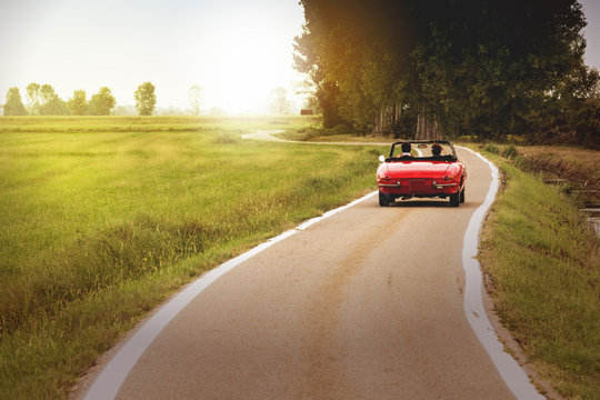 Fototapeta Klasyczny czerwony samochód kabriolet podróżujący po wsi o zachodzie słońca