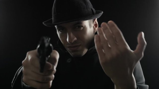 Portrait of a gangster in a hat and a black cloak threatens with weapon. Mafioso aiming a gun at the camera