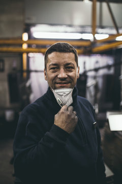 Industrial Manufacture. Dirty And Smiling Hard Job Worker Portrait. He Standing, Holding Protective Mask And Looking At Camera. Extreme Low Light Conditions And Noise Visible.