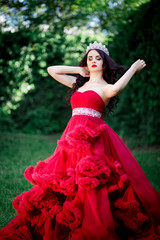 Fototapeta premium beautiful brunette in a crown with a bouquet in his hand and in a chic big and lush red dress in the park among the green