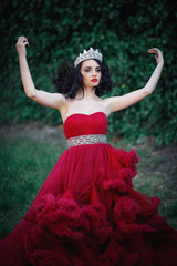 Fototapeta premium beautiful brunette in a crown with a bouquet in his hand and in a chic big and lush red dress in the park among the green