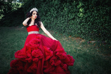 Fototapeta premium beautiful brunette in a crown with a bouquet in his hand and in a chic big and lush red dress in the park among the green