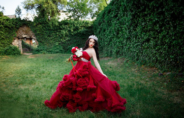 beautiful brunette in a crown with a bouquet in his hand and in a chic big and lush red dress in the park among the green