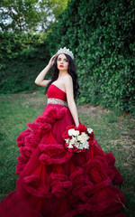 Obraz na płótnie Canvas beautiful brunette in a crown with a bouquet in his hand and in a chic big and lush red dress in the park among the green
