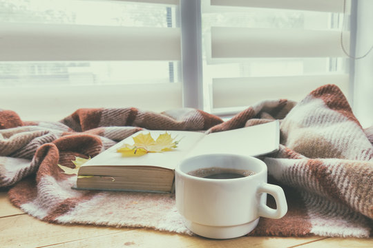 Cup of coffee and book with autumn yellow leaf surrounded wool blanket on windowsill