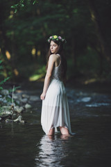 Artistic outdoor portrait of beautiful young woman wearing white dress and floral wreath on her head. Girl walking alone through cold mountain river.