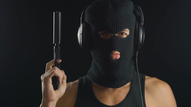man in a balaclava and A-shirt with gun listening to music on headphones