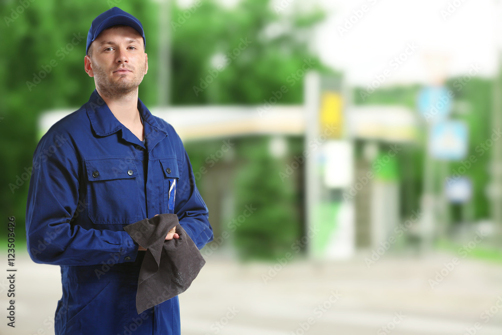 Wall mural Young mechanic in uniform with rag standing on blurred petrol station