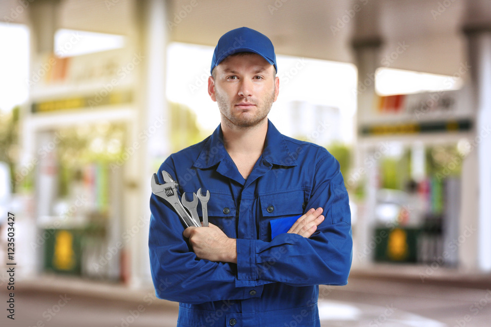 Wall mural Young mechanic in uniform with crossed arms and wrenches standing on blurred petrol station