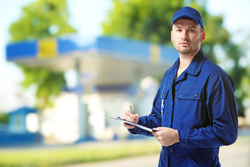 Young mechanic in uniform with a clipboard and pen on gas station blurred background