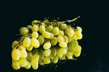 A branch of green grapes with large drops of water lies on a black background and reflects in the mirror. Background