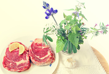 Raw beef steak with spices (sliced lemon and garlic) ready for cooking. Wild herbs and flowers on the wood board. Food ingredients and herbs still life. Toned colors vintage image