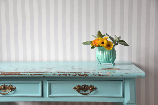 Ett turkost gammaldags skrivbord. En gullig pastellfärgad vas med Ringblommor på bordet.  Randiga tapeter på väggen.  Färgglad inredning och dekoration. 