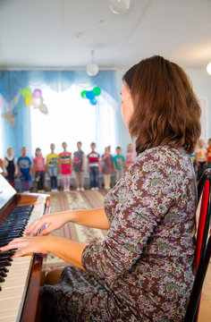 a young woman plays the piano for children , kids sing.